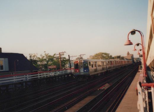 A silver and blue CTA 'L' train with the number 2504 is stopped at an elevated station platform with a red sign that reads Wellington.