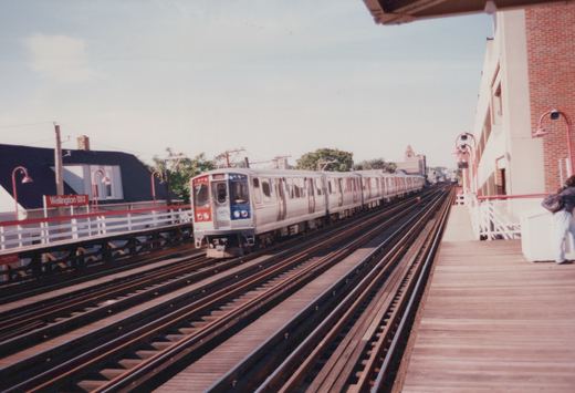 A silver, red and blue CTA train with the number 2475 is stopped at the Wellington elevated station platform under a clear sky.