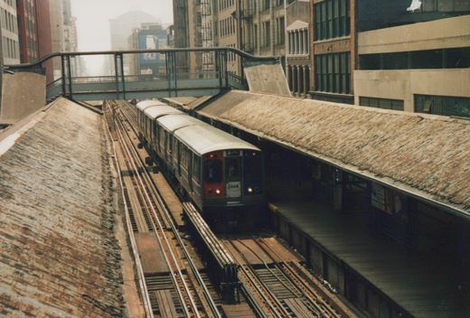 An elevated train with the number 2468 on its front, stopped at a station with a rusty roof, in a city setting.