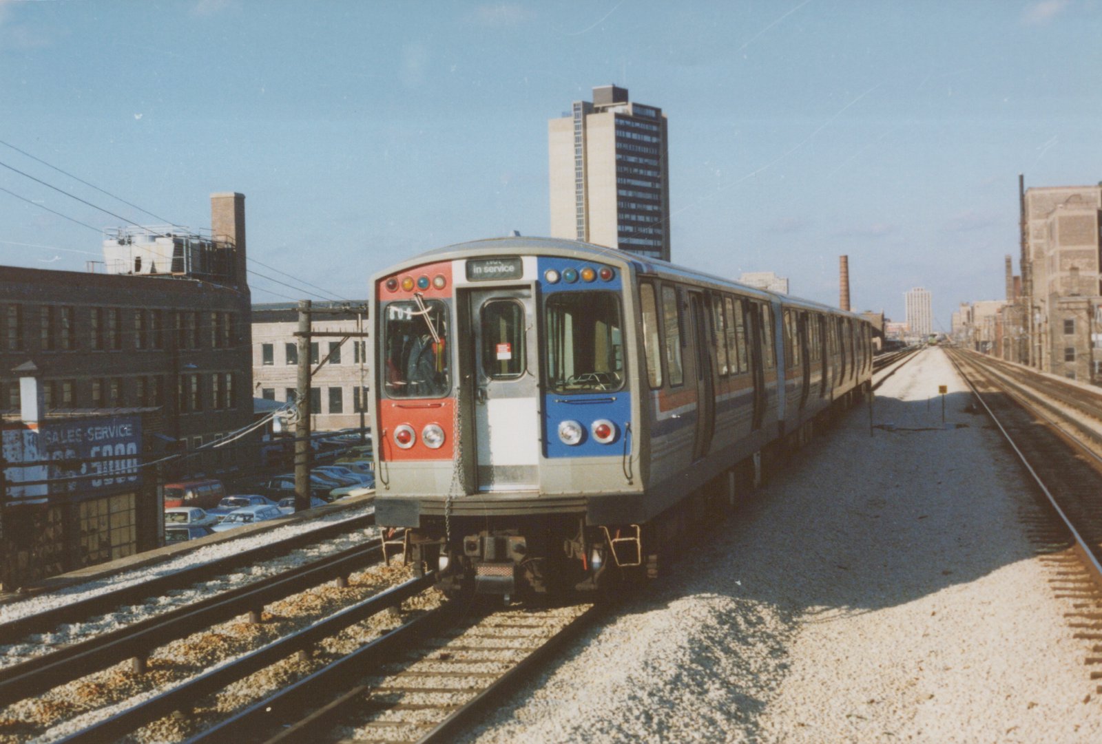 A silver CTA train car with red and blue front panels, numbered 802, sits on an elevated track with city buildings in the background under a clear blue sky.