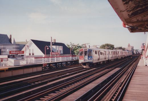A silver, red, and blue CTA train with the number 2430 is stopped at the elevated Wellington station on a sunny day with clear skies.