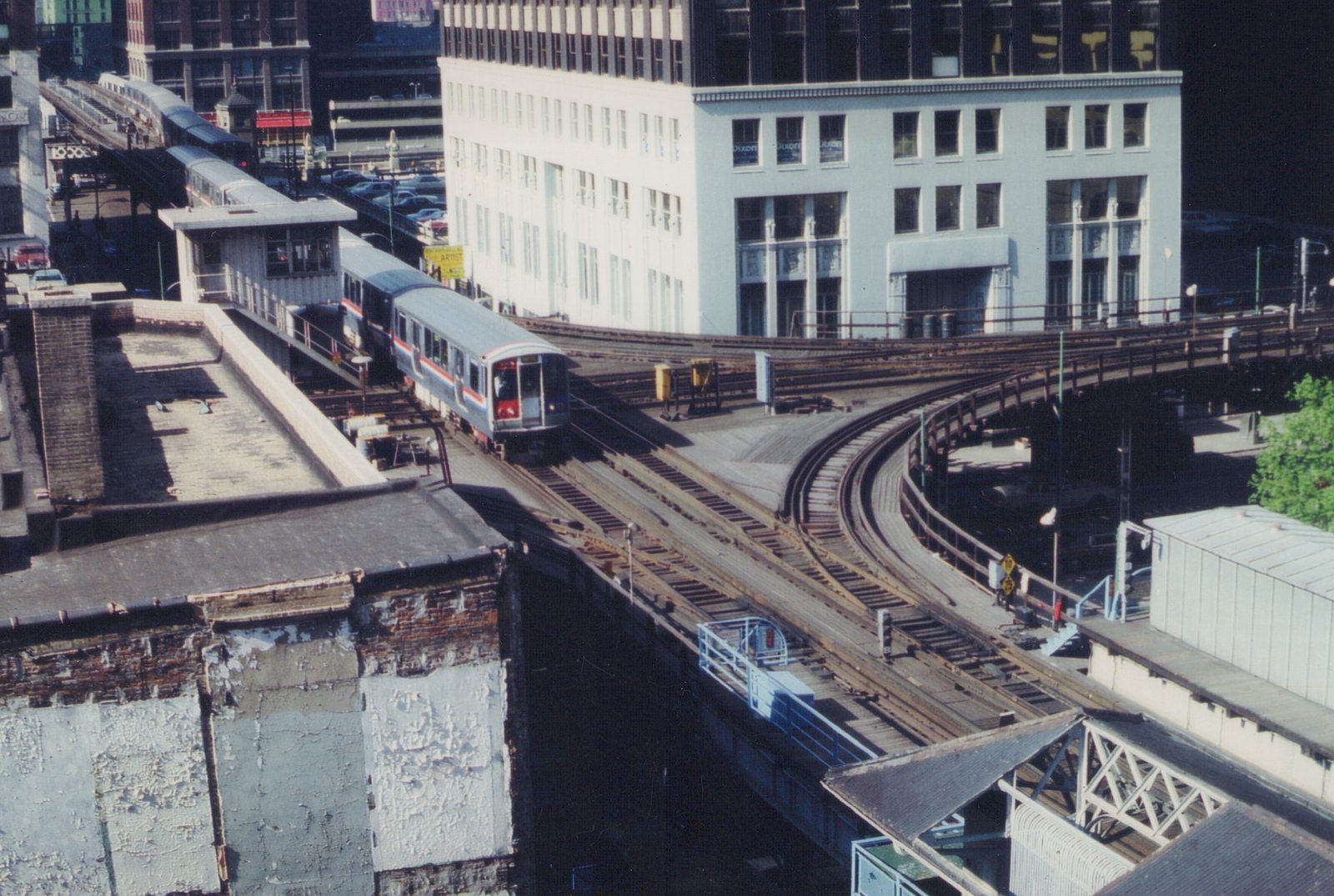 A silver CTA train with red and blue stripes travels along elevated tracks in downtown Chicago, passing a white building and a station platform.