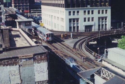 A silver CTA train with red and blue stripes travels along elevated tracks in downtown Chicago, passing a white building and a station platform.