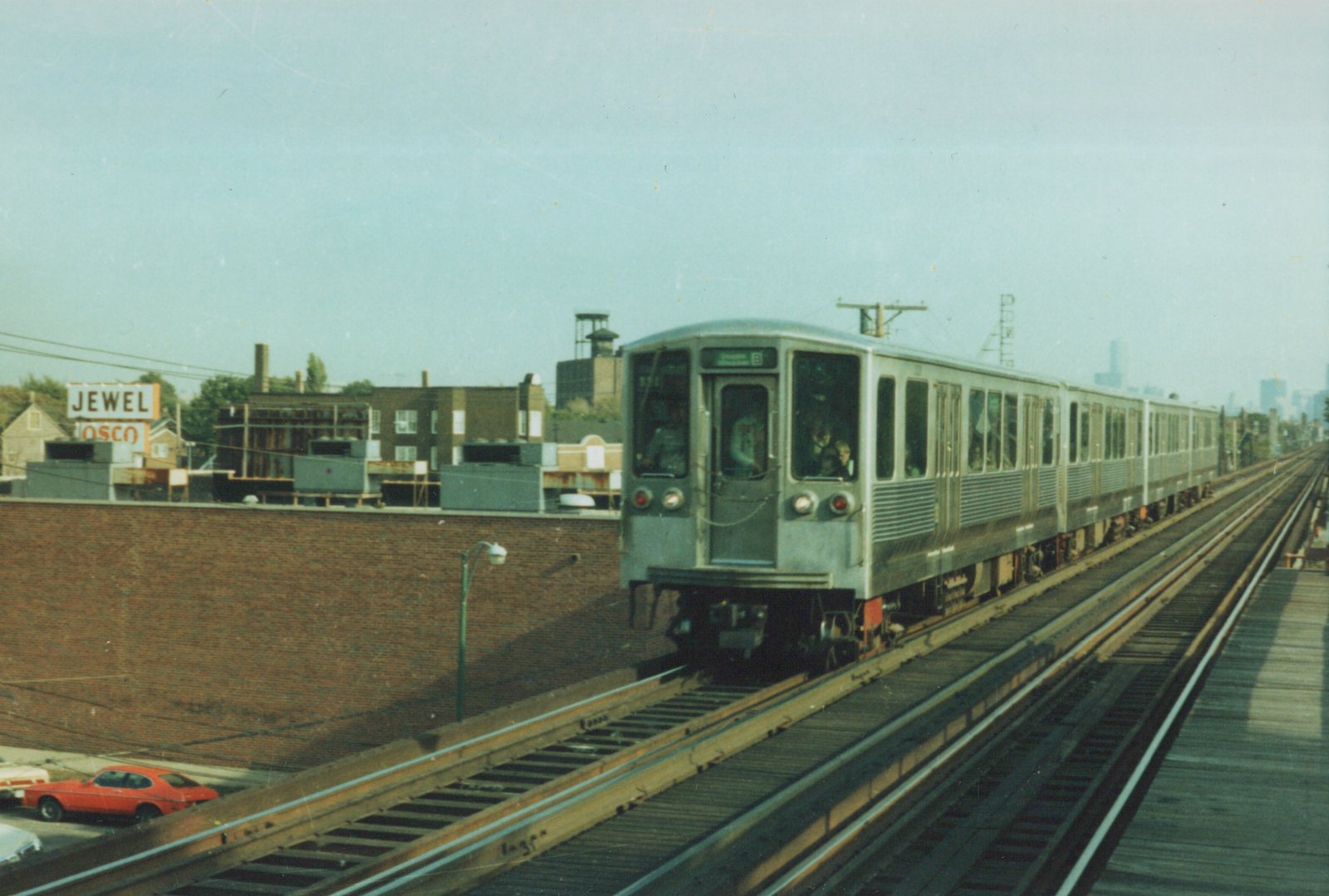 A silver Chicago Transit Authority train with the number 571 and destination 'Skokie' on its front, running on an elevated track with a brick wall and a Jewel-Osco sign in the background.