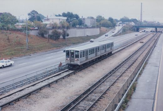 A silver two-car train moves along tracks in a highway median, with a police car and other vehicles on the road beside it, under a gray sky.