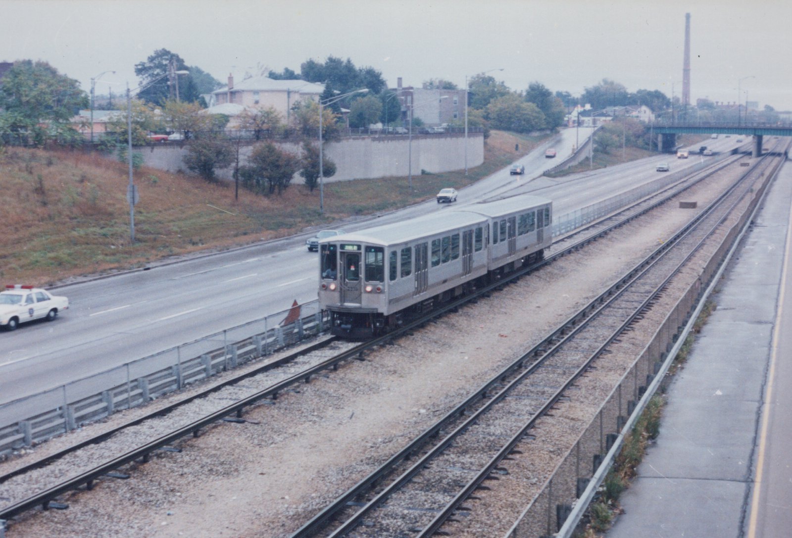 A silver two-car train moves along tracks in a highway median, with a police car and other vehicles on the road beside it, under a gray sky.