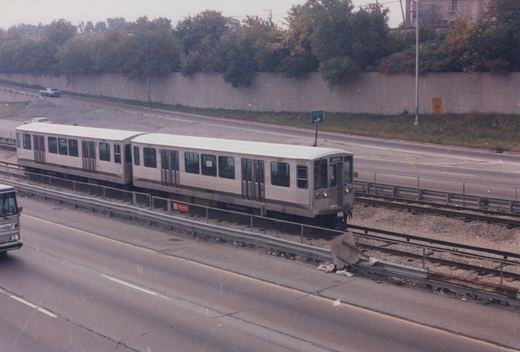 A silver CTA train with the number 2208 written on its side runs along tracks in the center of an expressway, with a green 'EXIT' sign and a yellow 'RAMP 30 MPH' sign visible in the background.