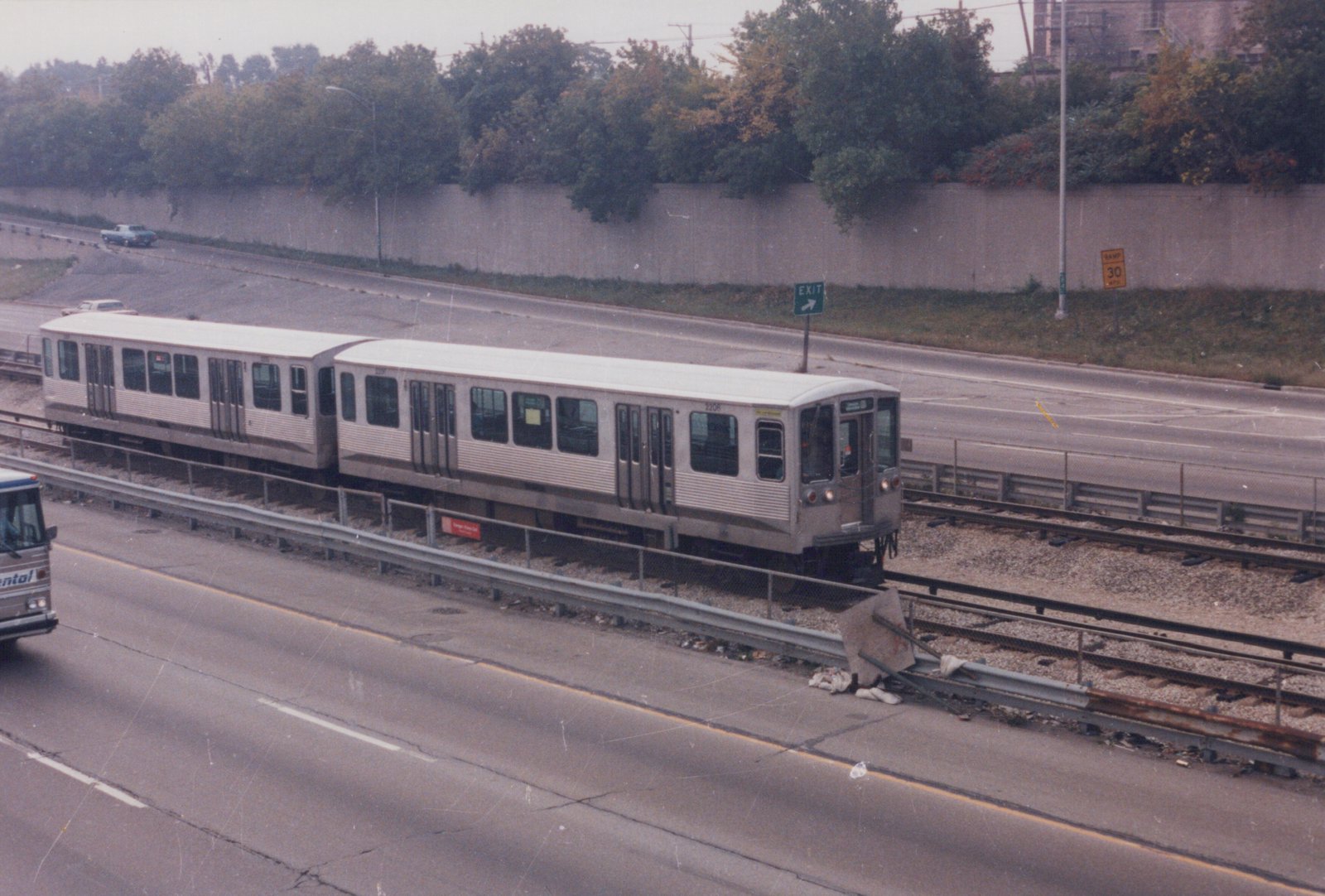 A silver CTA train with the number 2208 written on its side runs along tracks in the center of an expressway, with a green 'EXIT' sign and a yellow 'RAMP 30 MPH' sign visible in the background.