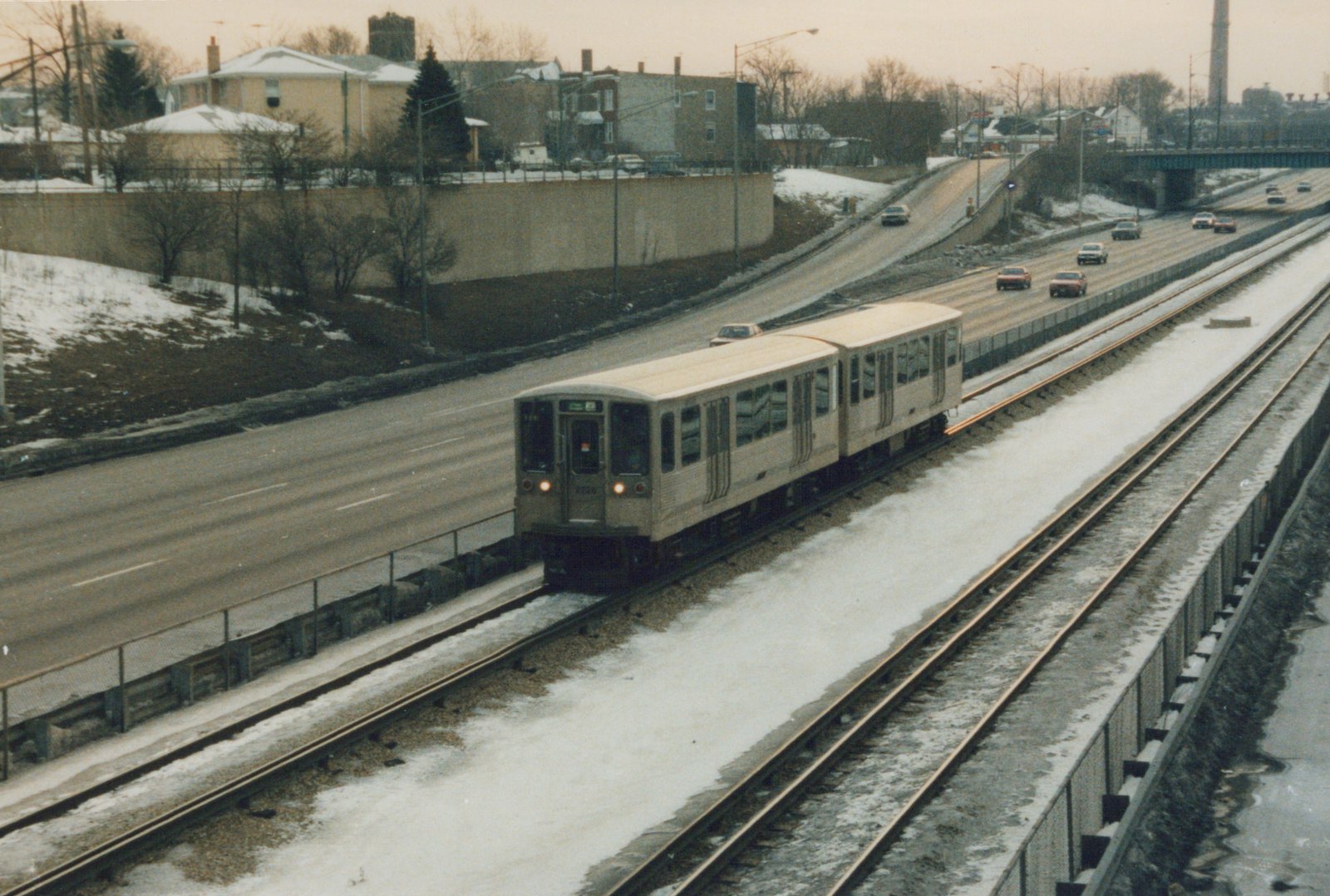 An elevated view of a two-car train of silver Chicago 'L' cars, number 2226 leading, marked for O'Hare, traveling on snow-dusted tracks in the median of a multi-lane highway.