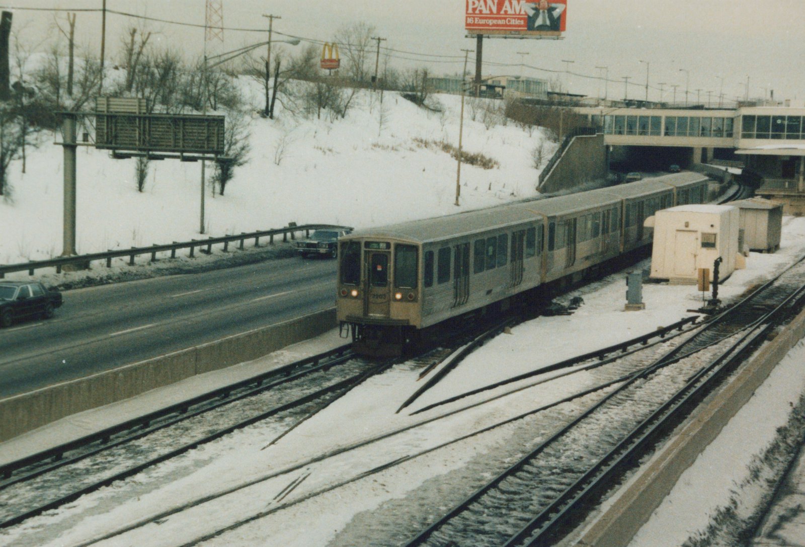 A four-car train of stainless steel rapid transit cars with a tan front, numbered 2203, runs on elevated tracks next to a snowy highway under an overcast sky.