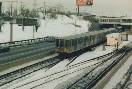 A four-car train of stainless steel rapid transit cars with a tan front, numbered 2203, runs on elevated tracks next to a snowy highway under an overcast sky.