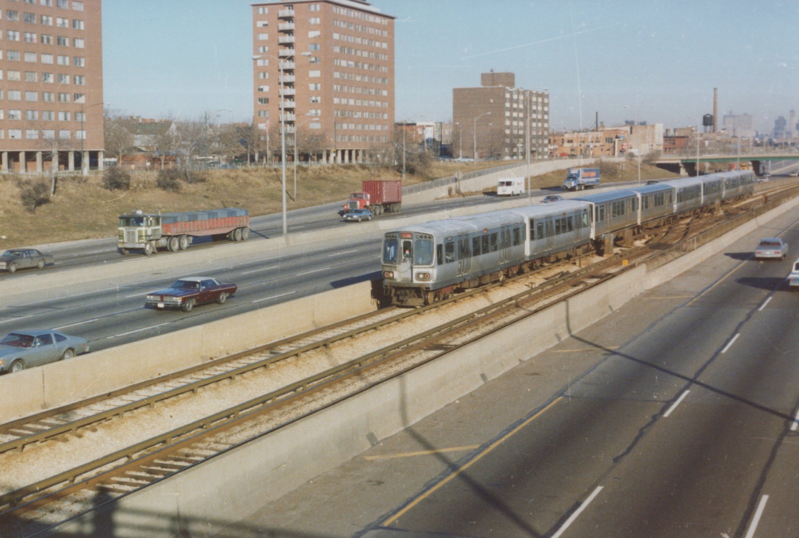 A silver Chicago 'L' train with the number 32 visible moves along elevated tracks between highway lanes, with brick apartment buildings in the background and vintage cars on the road.