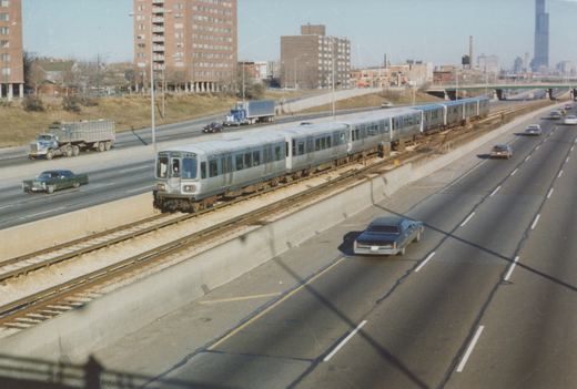 A silver Chicago 'L' train with a blue stripe moves along a highway median, surrounded by cars and trucks from the 1970s, with city buildings in the background.