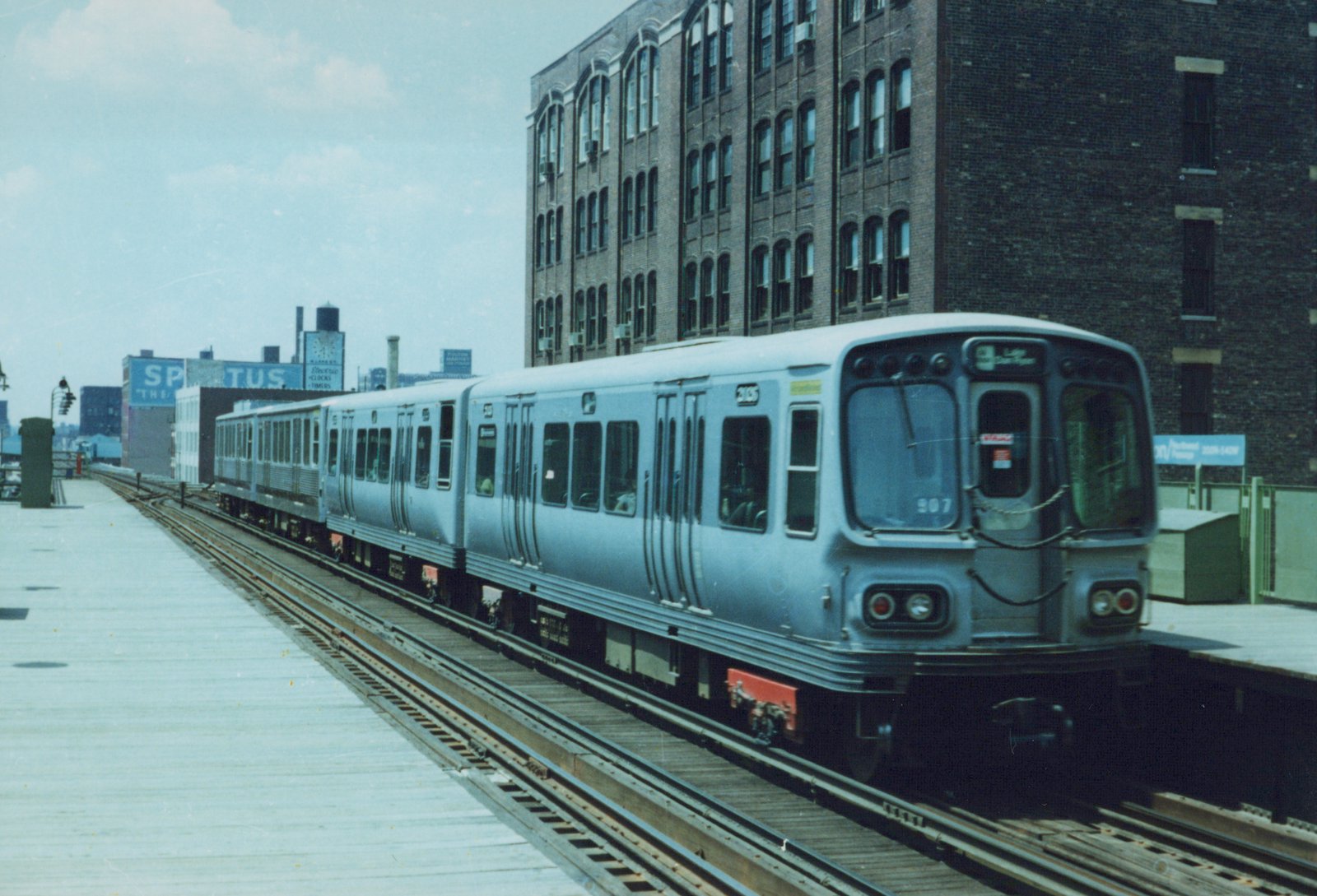 A silver rapid transit train with black front and red underframe is stopped at an elevated station; the lead car displays number 907 and destination 9 - LAKESHORE.