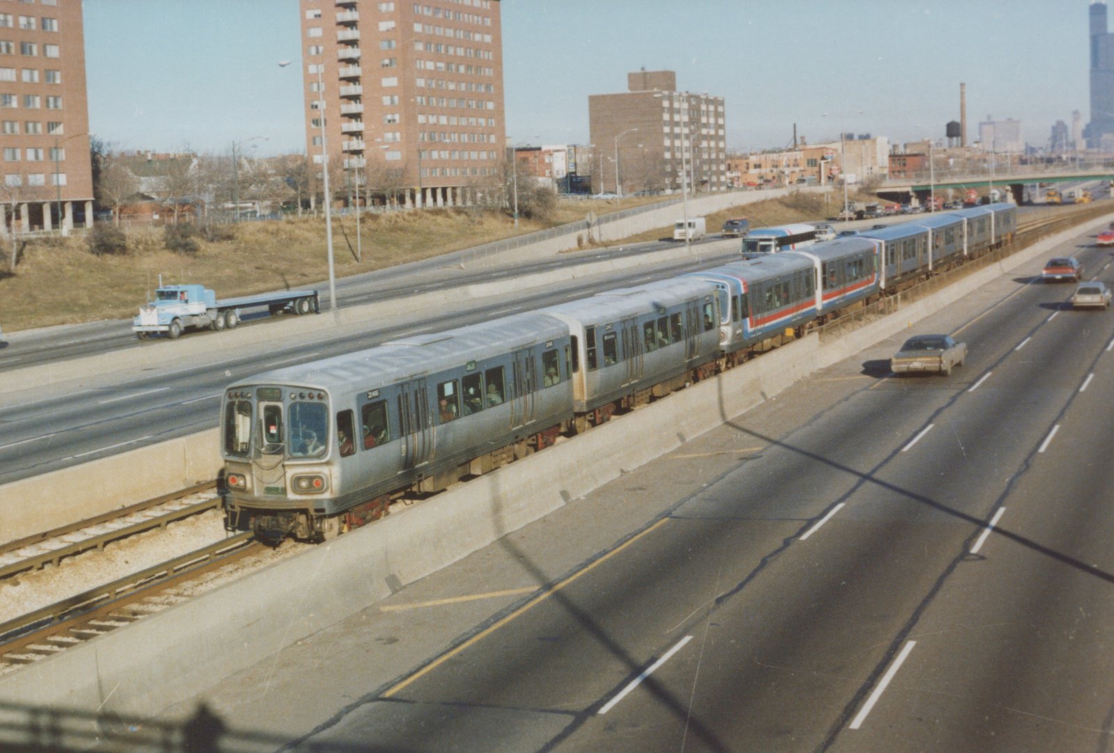 A silver CTA rapid transit train with red and blue stripes travels on tracks next to a multi-lane highway, with apartment buildings and a city skyline in the background.