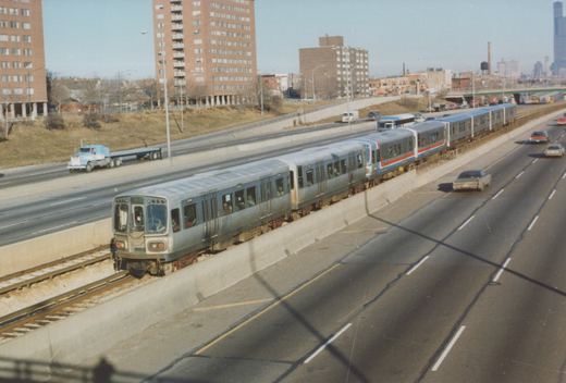 A silver CTA rapid transit train with red and blue stripes travels on tracks next to a multi-lane highway, with apartment buildings and a city skyline in the background.