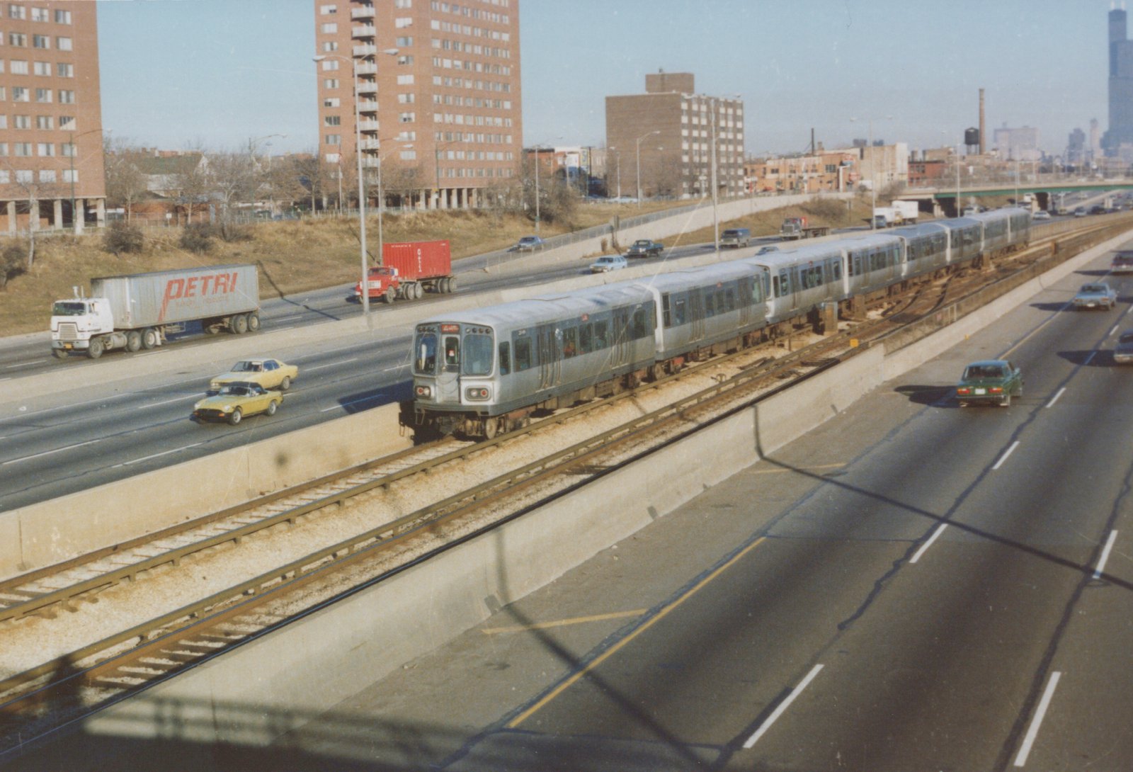 A silver CTA train with red and blue stripes travels on an elevated track next to a highway with cars and trucks, with brick apartment buildings in the background.