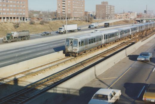A silver CTA rapid transit train numbered 3302 moves along an elevated track next to a highway with multiple cars and trucks, set against a backdrop of brick buildings and a clear winter sky.