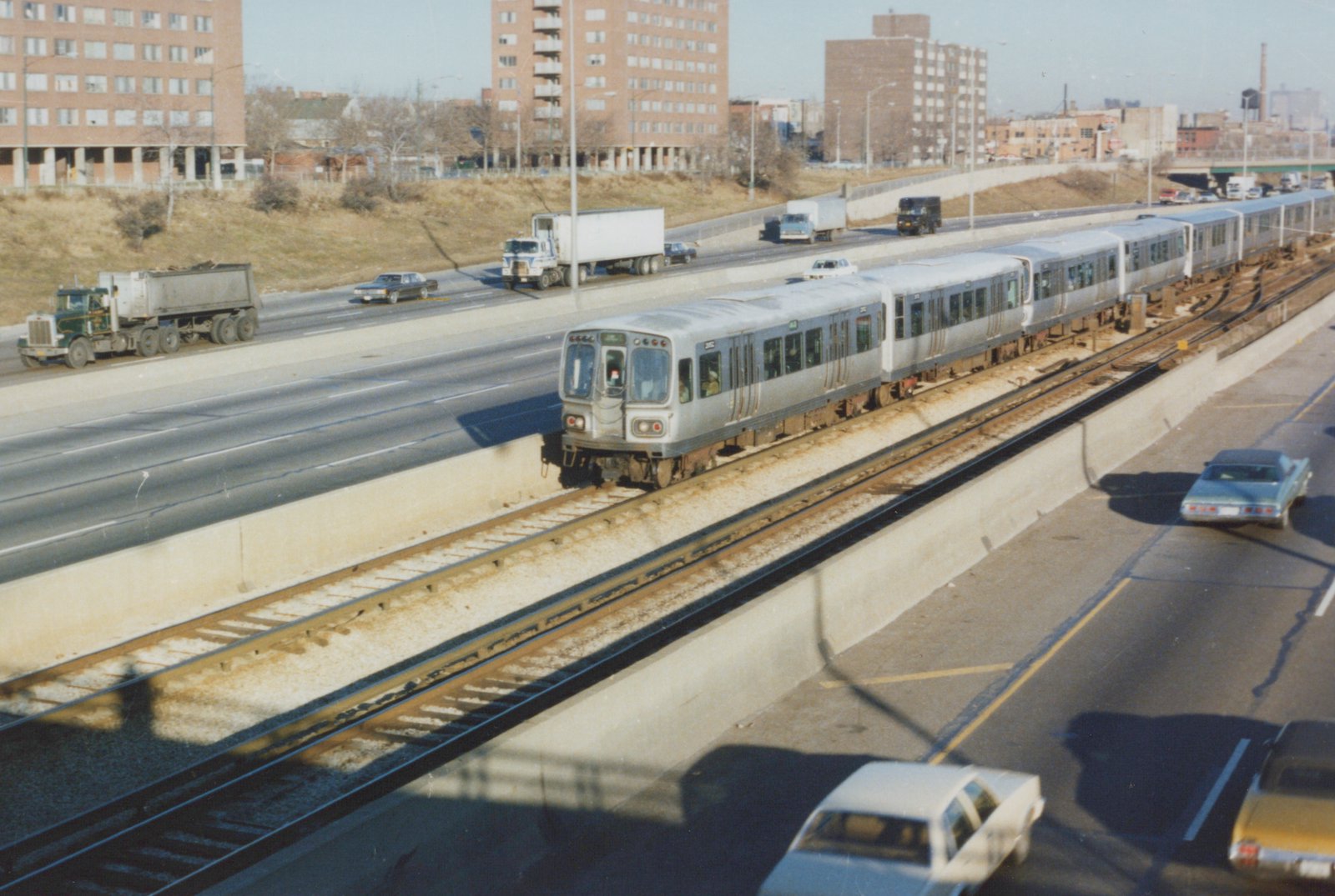 A silver CTA rapid transit train numbered 3302 moves along an elevated track next to a highway with multiple cars and trucks, set against a backdrop of brick buildings and a clear winter sky.