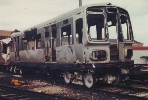 The burned-out shell of a CTA 2000-series rapid transit car sits on tracks in a scrap yard, with another similar car behind it, under an overcast sky.