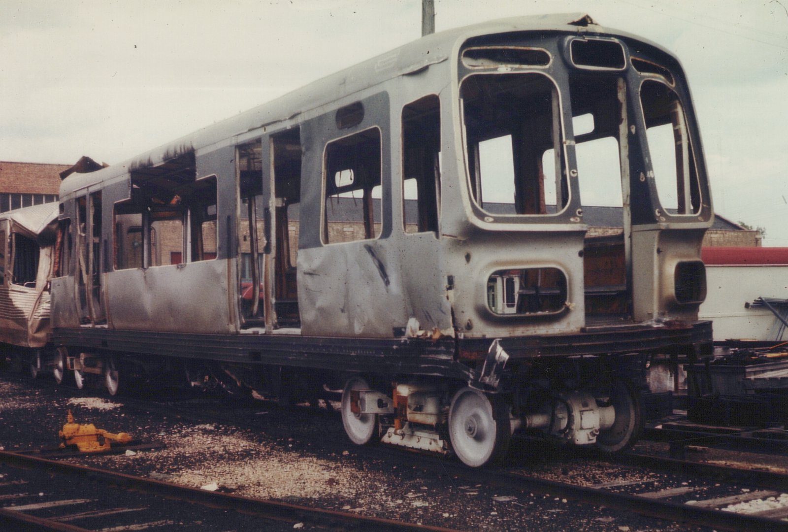 The burned-out shell of a CTA 2000-series rapid transit car sits on tracks in a scrap yard, with another similar car behind it, under an overcast sky.