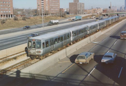 A silver CTA rapid transit train with the number 2040 on its side moves along an elevated track next to a multi-lane highway with several vintage cars from the 1970s.