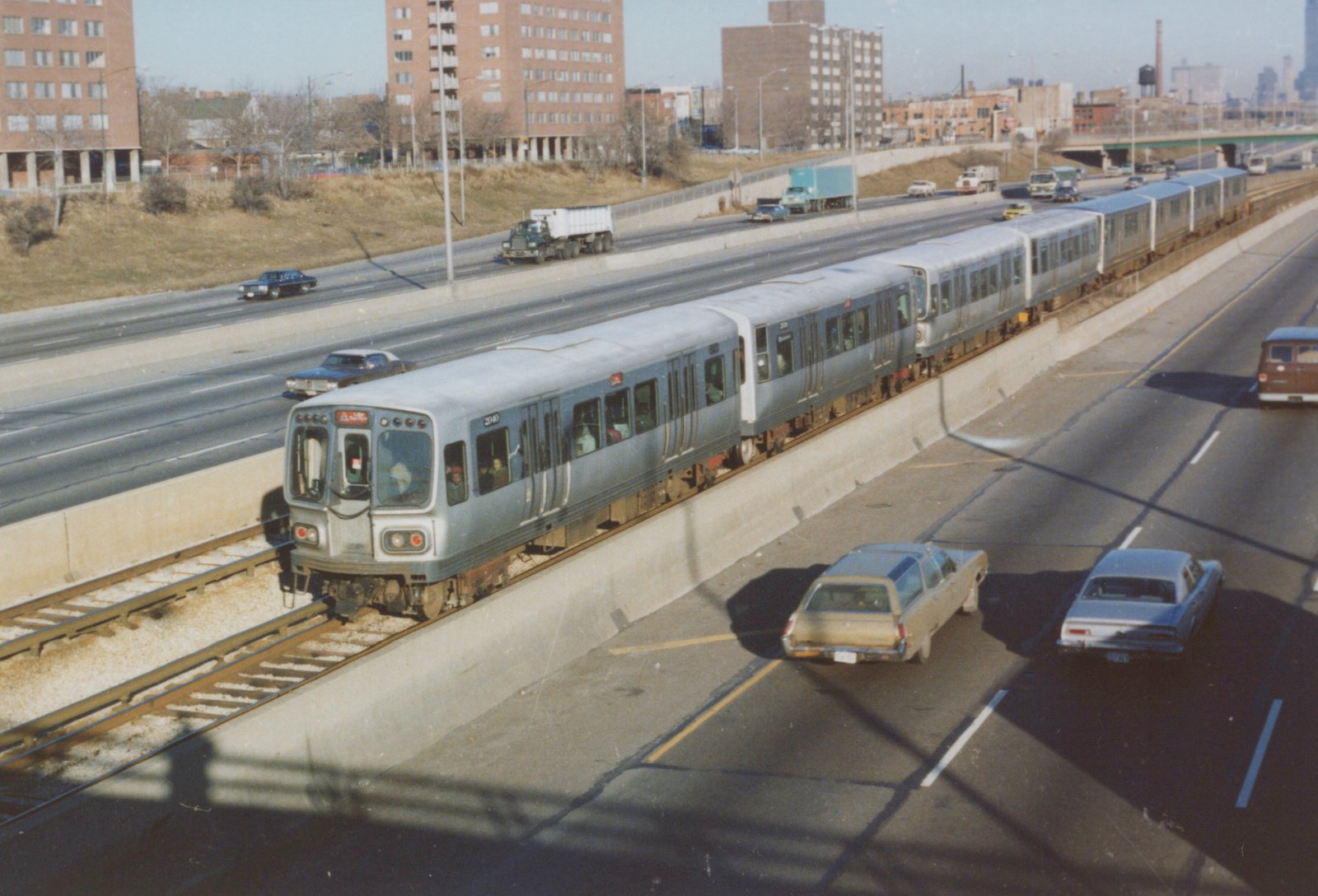 A silver CTA rapid transit train with the number 2040 on its side moves along an elevated track next to a multi-lane highway with several vintage cars from the 1970s.