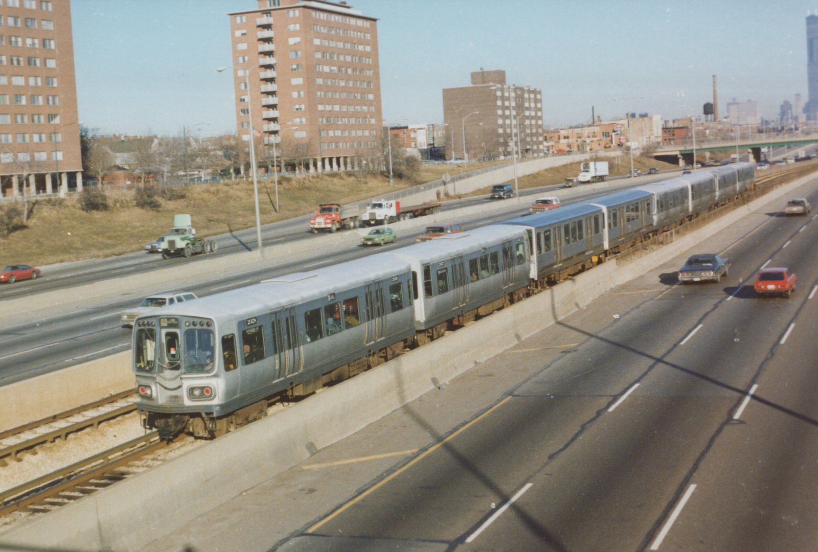 A silver and blue Chicago 'L' train travels along an expressway median with cars driving on the highway beside it.