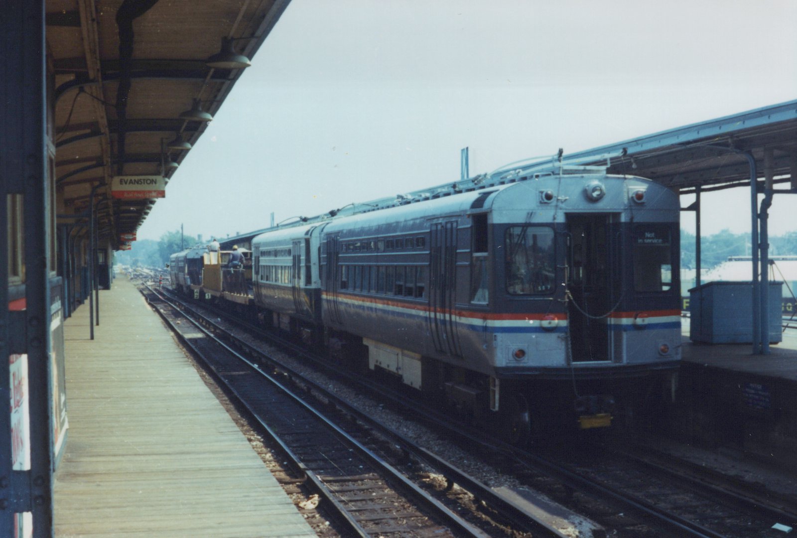 A silver electric train with a red and blue stripe, marked 'Not in service,' is stopped at a station platform with a wooden work car and two workers behind it. A sign reads 'EVANSTON.'