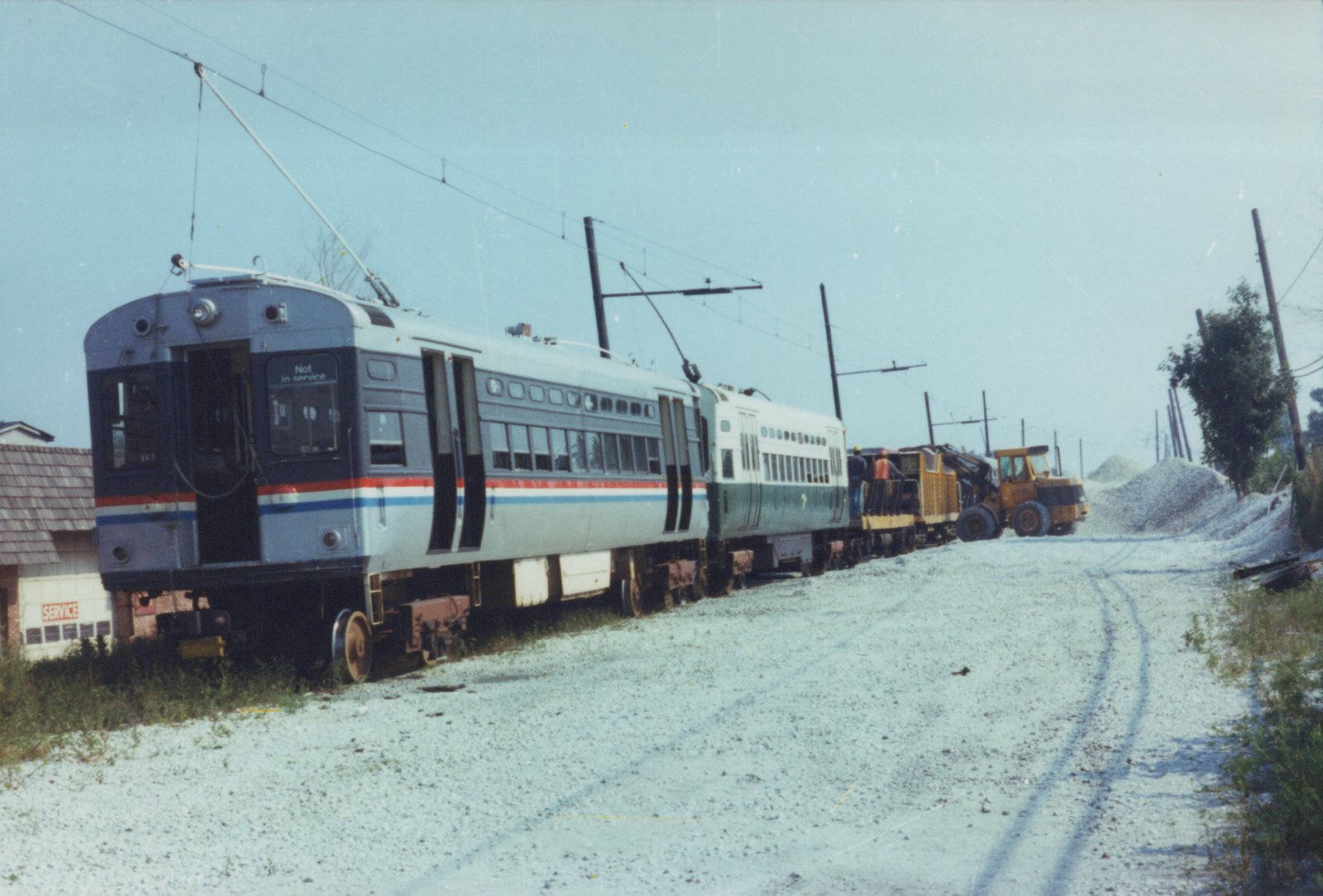 Two silver Amtrak Metroliner trains with red and blue stripes sit on a gravel track under wires, with a yellow construction vehicle and two people on the rear platform.