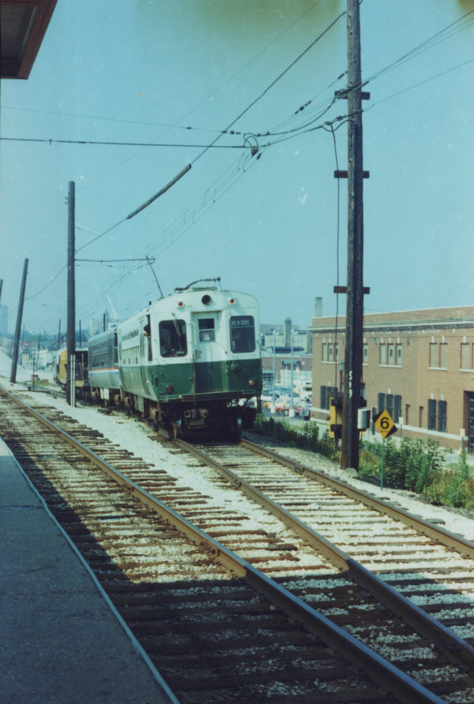 A green and cream South Shore Line train car, number 80, with a 'NOT IN SERVICE' sign, is moving along tracks beside a street lined with buildings and parked cars under a clear sky.