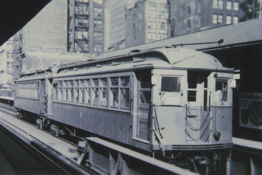A black and white photo of a vintage wooden 'L' car with a clerestory roof, stopped at an elevated station. The front sign reads 'A LAKE', and a 'Del Monte Fruit Co.' ad is visible on the platform wall.