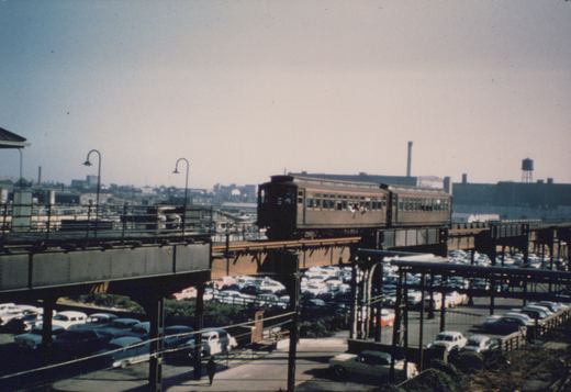 A vintage brown and cream Chicago transit train runs on an elevated track above a street lined with classic 1950s cars and a lone pedestrian.