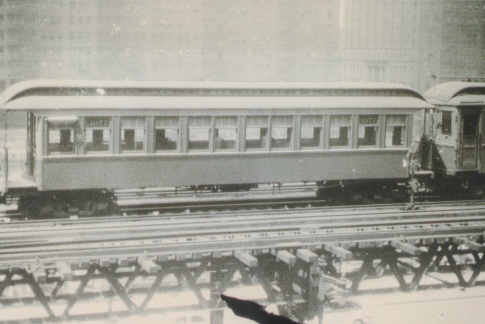 A black-and-white photo of a single, old rapid transit car, number 2077, on an elevated track with snow on the ground and city buildings in the background.