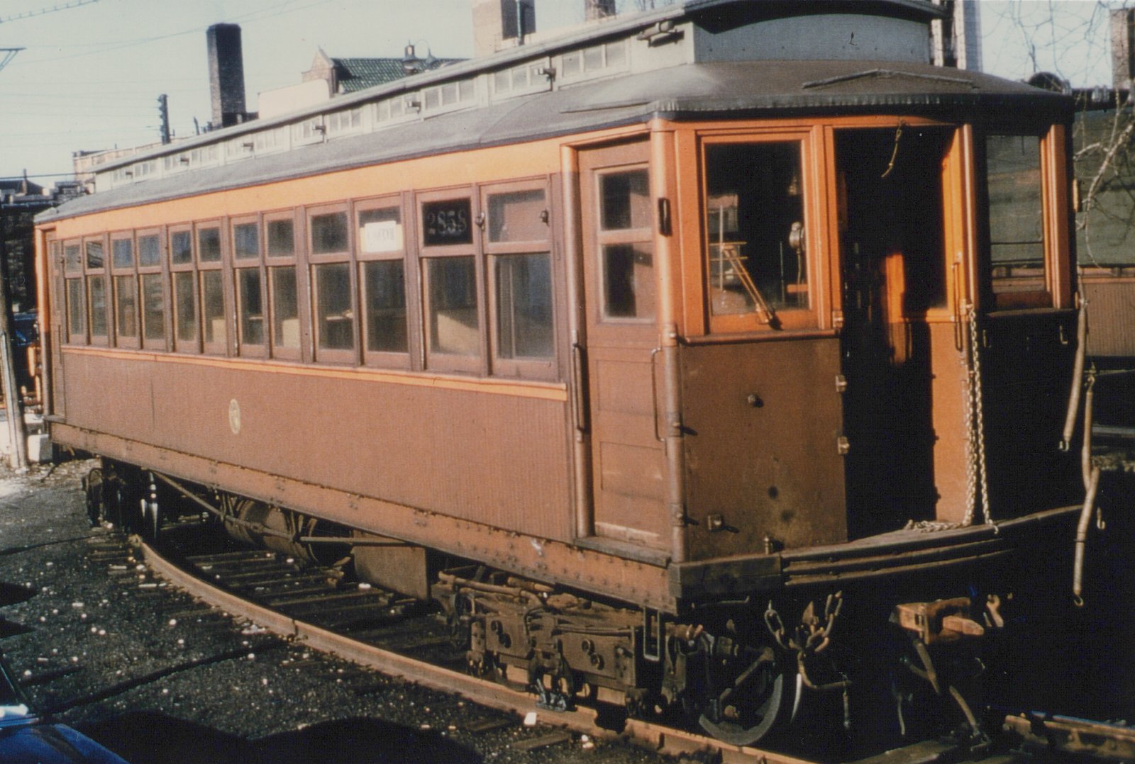 A three-quarter rear view of a vintage brown and yellow interurban car, numbered 2858, sitting on tracks in a yard with a building and a blue car visible in the background.