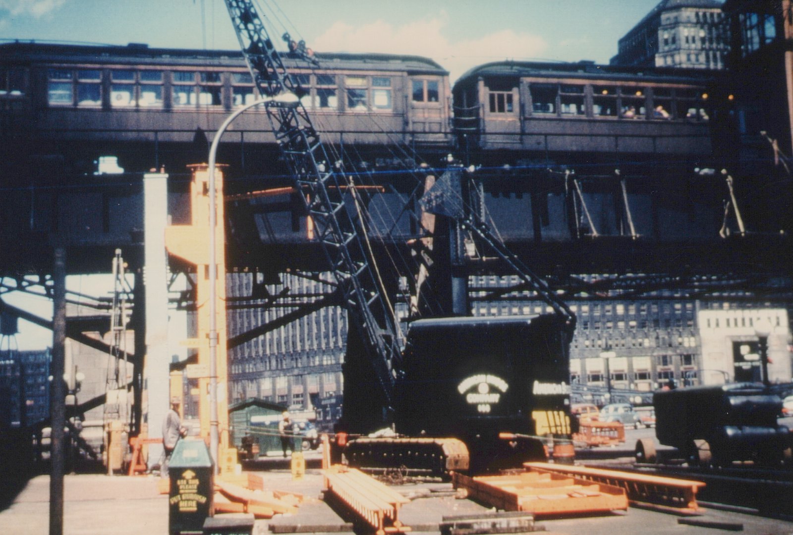 Two dark green and cream rapid transit cars travel on an elevated steel trestle above a street scene with a large black steam crane and several vintage automobiles.