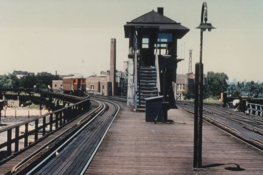 A maroon and cream rapid transit car on a wooden elevated track with a tower and brick buildings in the background.
