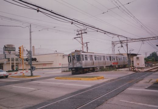 A silver rapid transit train with the number 311 is stopped at a street-level railroad crossing in a suburban area, with a lumber yard and traffic lights nearby.