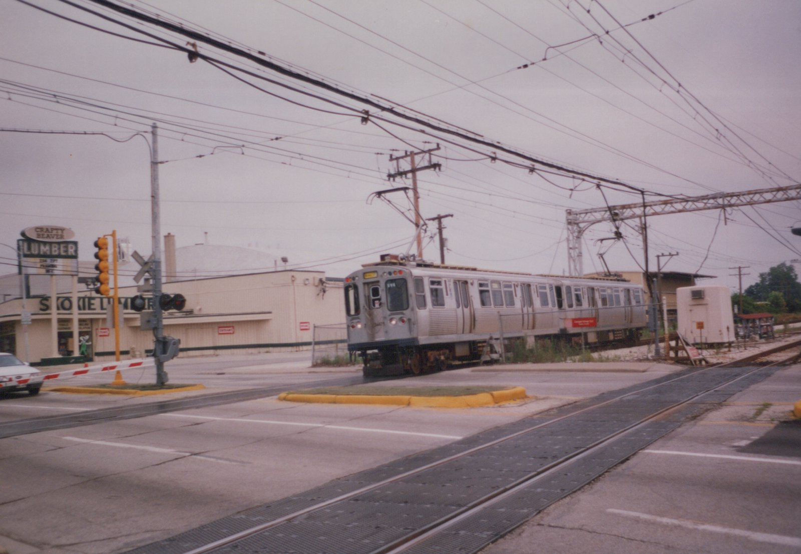 A silver rapid transit train with the number 311 is stopped at a street-level railroad crossing in a suburban area, with a lumber yard and traffic lights nearby.