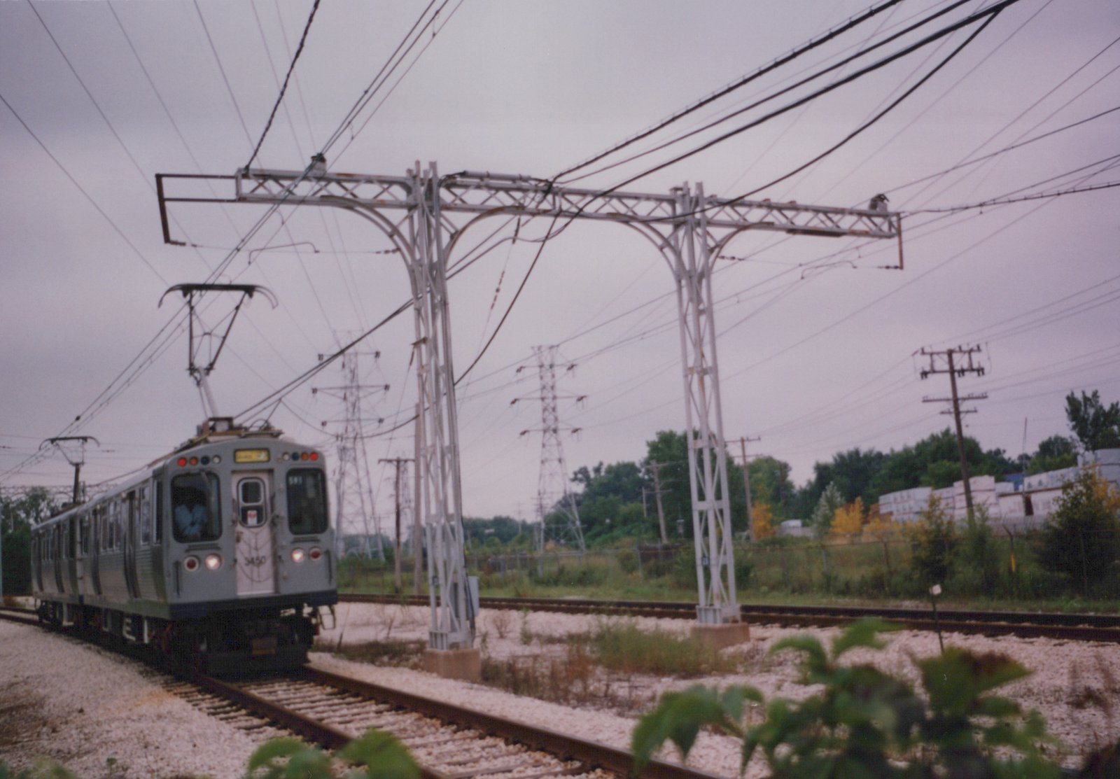 A gray rapid transit train with the number 3450 on its front moves along tracks under a complex web of overhead wires, with trees and power lines in the background under a gray sky.