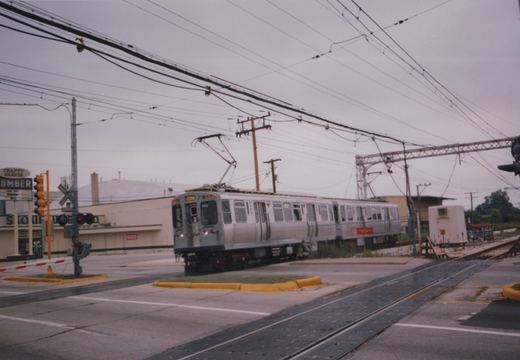 A silver CTA rapid transit car with the number 583 on its front is crossing a paved street at a grade crossing, with a lumber yard sign visible in the background under a gray sky.