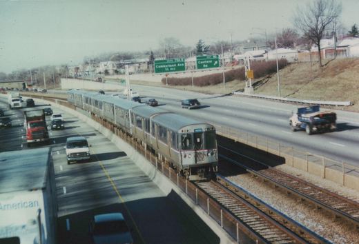 A silver CTA train with red, white, and blue stripes runs on tracks in the middle of a busy highway, surrounded by cars and trucks, with green highway signs for Cumberland Ave and Canfield Rd visible in the background.