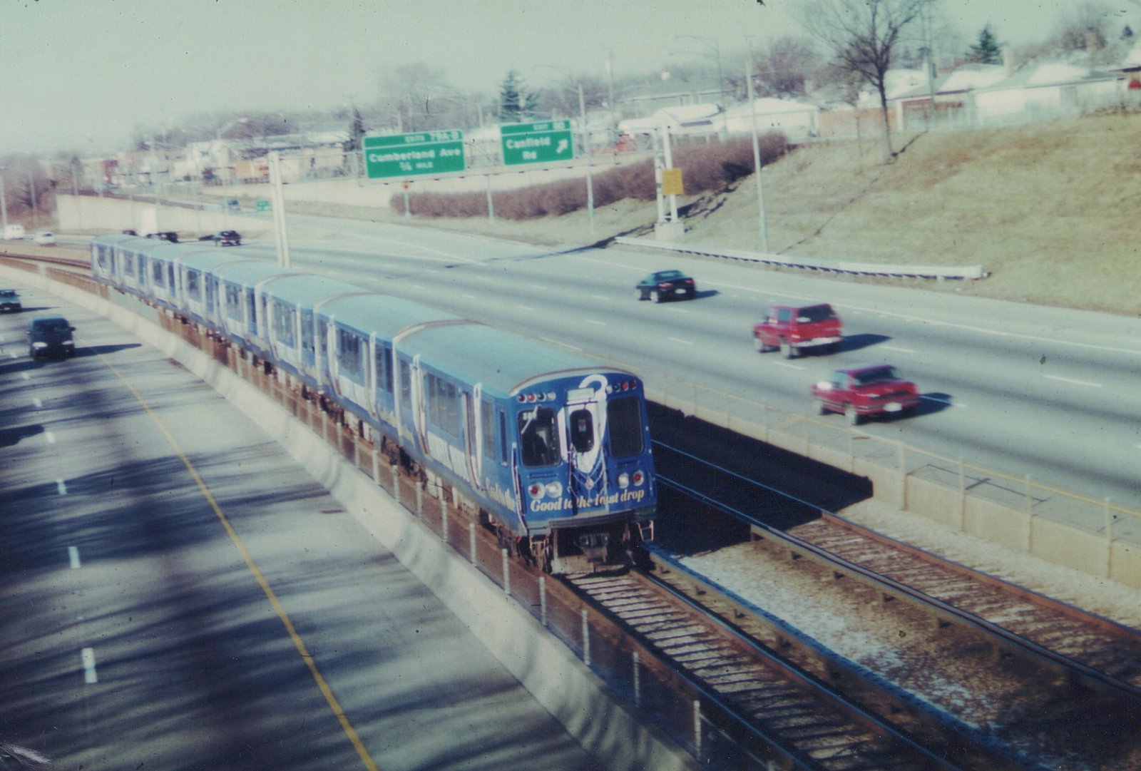 A blue CTA train on tracks in the middle of a highway, with cars driving on either side and highway signs visible in the background.