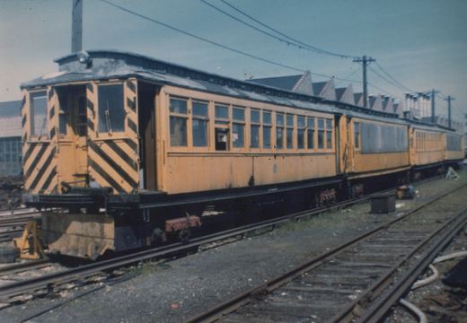 A row of yellow interurban railroad cars with black diagonal stripes on the front, parked on tracks in a yard under a clear blue sky.