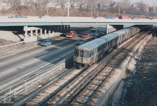 An elevated view of a silver Chicago 'L' train with red and blue stripes, numbered 221, running on tracks parallel to a multi-lane highway with cars and an overpass in the background on a clear winter day.