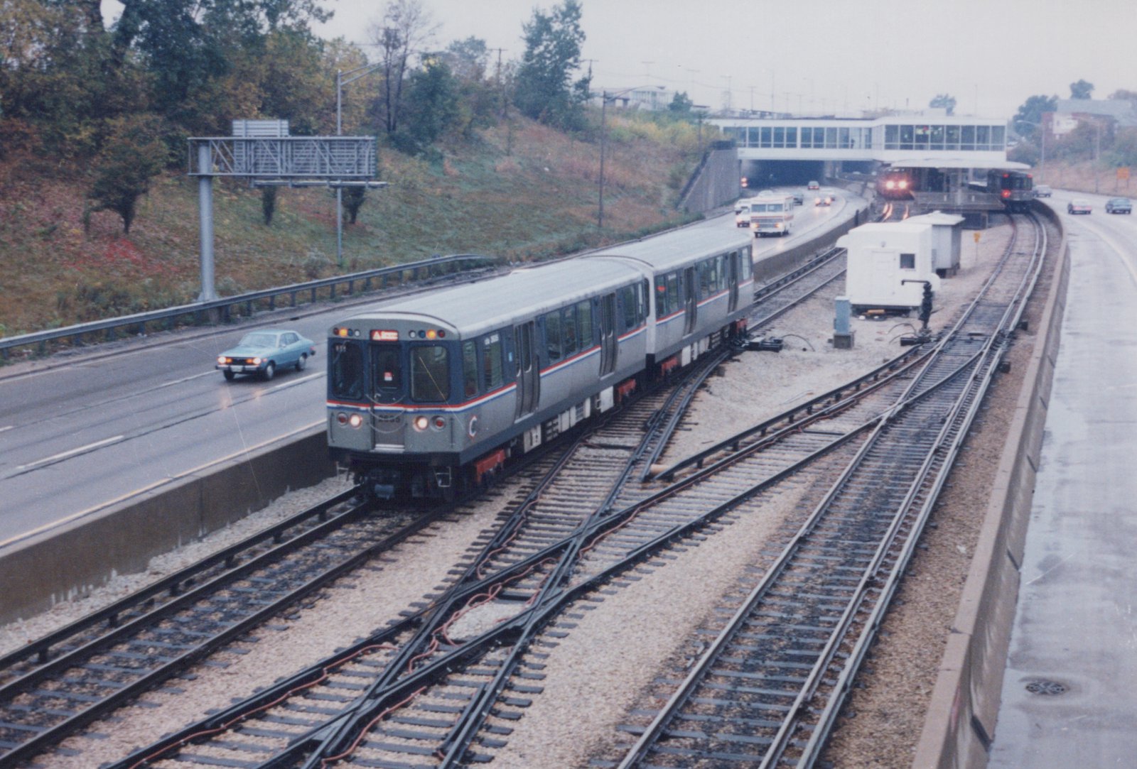 A silver and blue Chicago 'L' train travels on tracks in the median of a wet highway, with cars and a station visible nearby on an overcast autumn day.