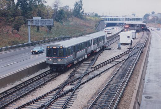 A silver and blue Chicago 'L' train travels on tracks in the median of a wet highway, with cars and a station visible nearby on an overcast autumn day.