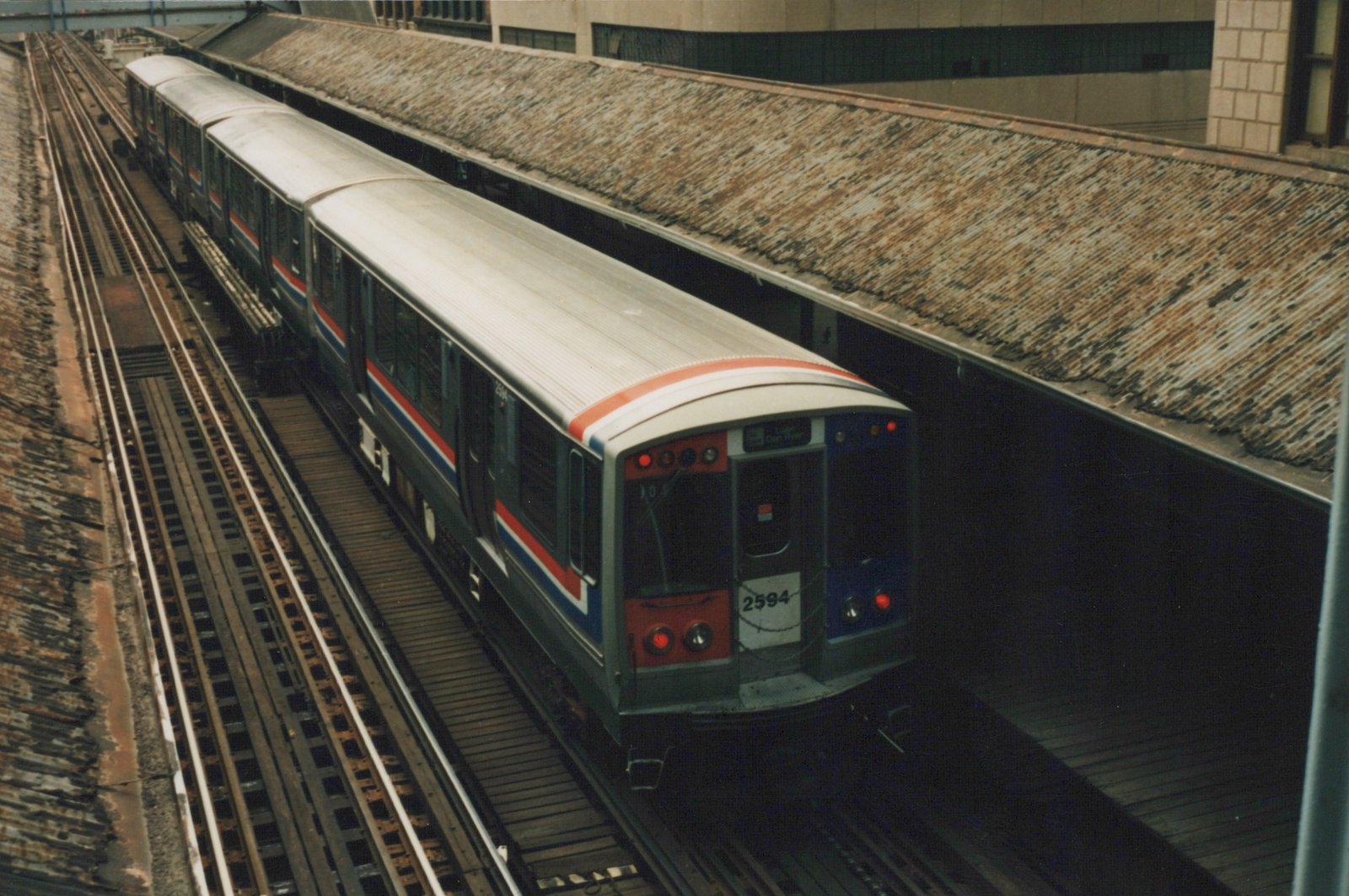 A high-angle view of a Chicago Transit Authority 2600 series rapid transit train, car number 2594, stopped at an elevated station with a rusty roof. The destination sign reads 'LAKE + DAN RYAN'.