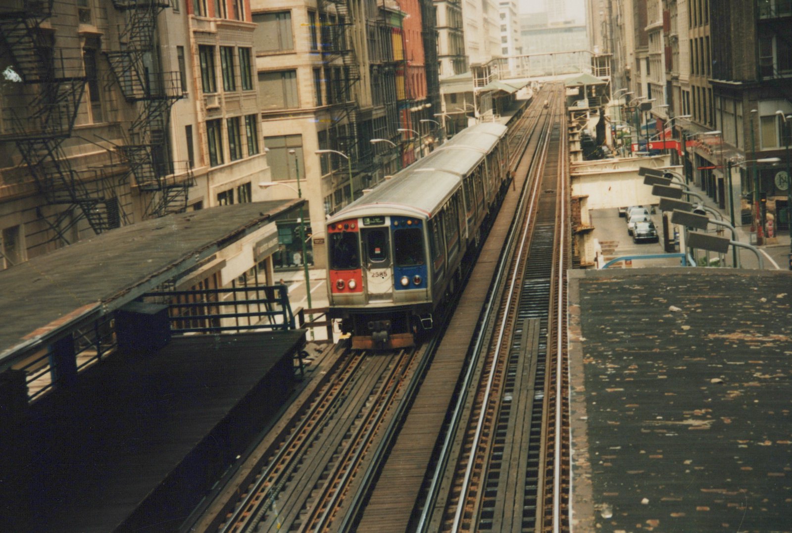 A red, white, and blue Chicago 'L' train marked '2585' travels on an elevated track between buildings, with a platform visible on the left.