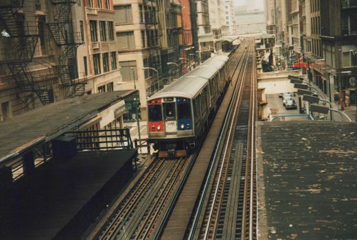 A red, white, and blue Chicago 'L' train marked '2585' travels on an elevated track between buildings, with a platform visible on the left.